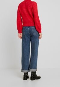 Roter gestrickter Pullover mit tailliertem Saum und Ballonärmeln, kombiniert mit hochgeschnittenen blauen Jeans, die an den Säumen aufgerollt sind. Schwarze Knöchelstiefel.