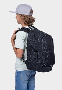 Czarny plecak w wzór nietoperzy, z wyściełanymi szelkami, wieloma zamykanymi na zamek kieszeniami i teksturowanym wykończeniem, noszony na jasnej koszuli.