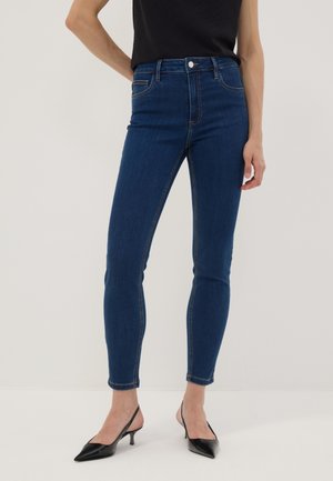 Frau trägt dunkelblaue Skinny Jeans und schwarze spitze Slingback-Pumps, steht vor einem schlichten hellen Hintergrund.