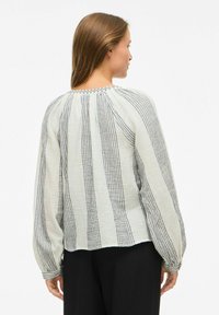 Femme avec de longs cheveux bruns portant un chemisier blanc lâche à rayures noires verticales et un pantalon noir, vue de dos.