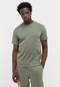 Młody mężczyzna z krótkimi, kręconymi włosami, ubrany w dopasowaną oliwkową koszulkę i dresy, stoi z jedną ręką w kieszeni na tle jednolitym.