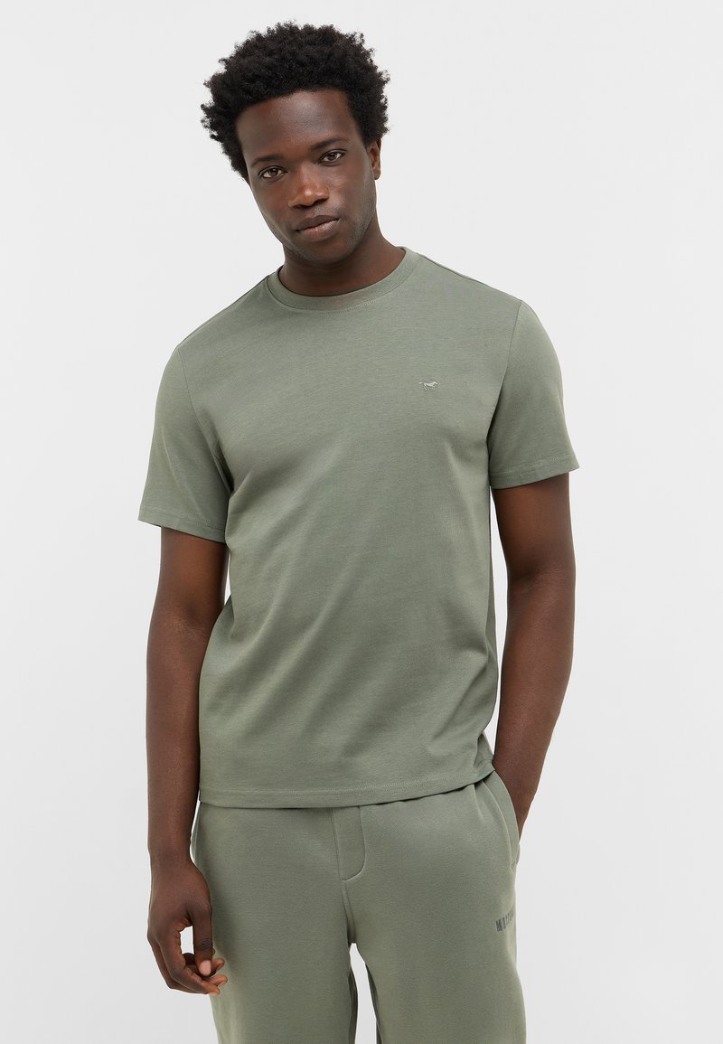 Młody mężczyzna z krótkimi, kręconymi włosami, ubrany w dopasowaną oliwkową koszulkę i dresy, stoi z jedną ręką w kieszeni na tle jednolitym.