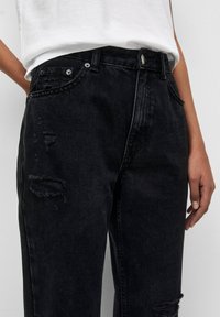 Černé džíny s vysokým pasem z denimové látky se zvýrazněnými prvky opotřebení, viditelným šitím a stříbrnými detaily. Tkanina má strukturovaný, drsný vzhled.