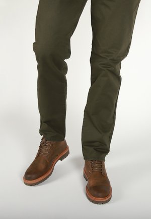Brown leather lace-up boots with a rounded toe, textured surface, and rugged rubber sole, paired with dark green trousers.