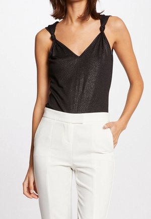 Vrouw draagt een mouwloze zwarte glinsterende top met geknoopte bandjes, gecombineerd met witte, op maat gemaakte, hooggetailleerde broek met zakken.