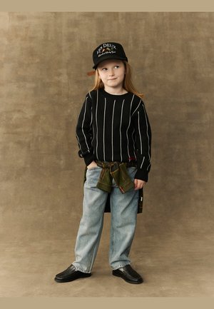 Svart strikkegenser med hvite striper, grønn rutete skjorte knyttet rundt livet, lyseblå jeans, svarte sko og en svart caps med logo.