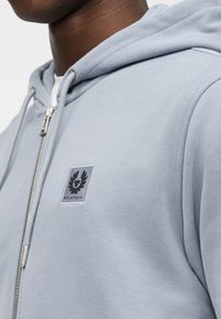 Κοντινό πλάνο ενός ατόμου που φοράει ένα ανοιχτό μπλε φούτερ με φερμουάρ και κουκούλα, με ένα ψηφιακό έμβλημα Belstaff στο στήθος.