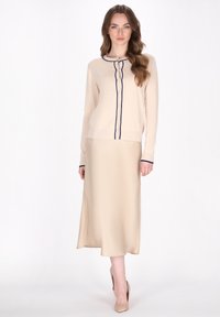 Beige cardigan met marineblauwe afwerking, knoopsluiting, gecombineerd met een beige satijnen midi-rok. Model draagt nude hakken, wat een slank silhouet benadrukt.