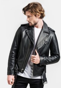 Schwarze Leder-Bikerjacke mit asymmetrischem Reißverschluss, Stehkragen und Druckknöpfen. Über einem weißen T-Shirt getragen, zeigt sie seitliche Taschen und ein Schnallen-Detail.