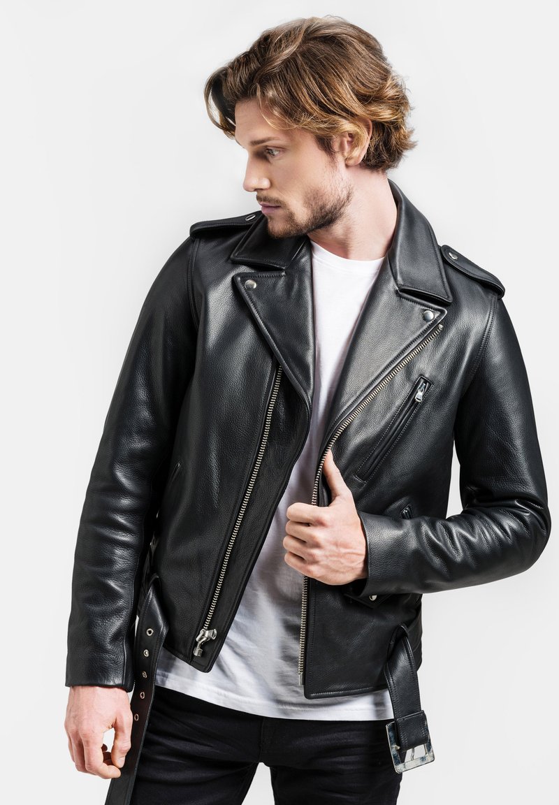 Schwarze Leder-Bikerjacke mit asymmetrischem Reißverschluss, Stehkragen und Druckknöpfen. Über einem weißen T-Shirt getragen, zeigt sie seitliche Taschen und ein Schnallen-Detail.