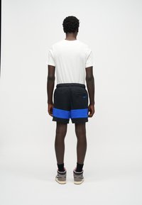 Άνδρας που στέκεται, φοράει λευκό μπλουζάκι, μαύρο σορτς με πλατιές μπλε ρίγες, μαύρες κάλτσες και γκρι παπούτσια τύπου high-top, σε απλό λευκό φόντο.