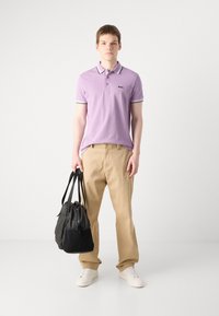 Polo majica u svijetloplavoj boji s kontrastnim bijelim ovratnikom i manžetama; bež hlače; crna torba za putovanje; bijele tenisice; model stoji.