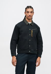 Νέος άνδρας με πλεξούδες φοράει μαύρο τζάκετ με κουμπιά και δερμάτινο γιακά, με τσέπη φερμουάρ, στέκεται μπροστά σε λευκό φόντο.