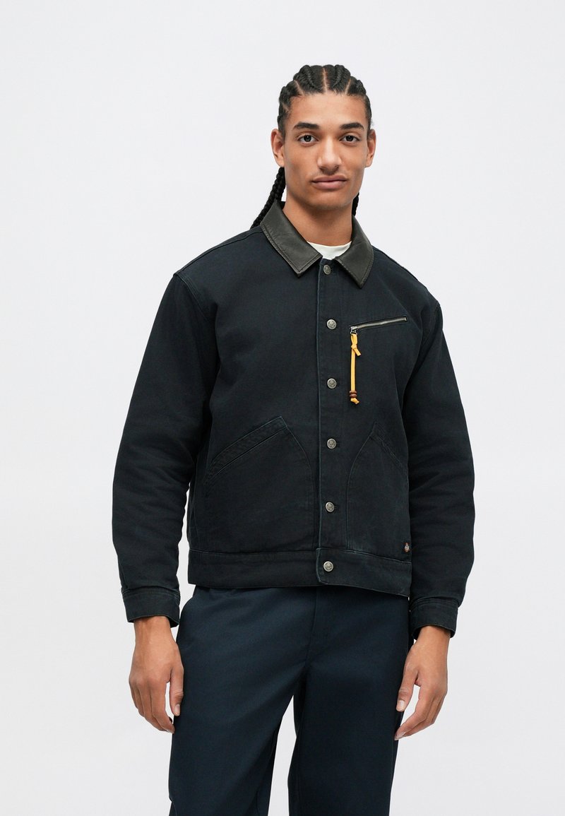 Young man with braided hair wears black buttoned jacket with leather collar and a zipper pocket, standing against a plain white background.