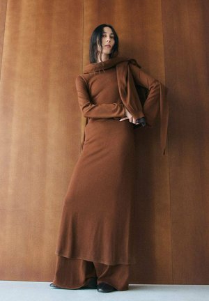 Vrouw in lange bruine jurk met bijpassend jasje over de schouder, staand tegen een houten wand met zichtbare zwarte schoenen.