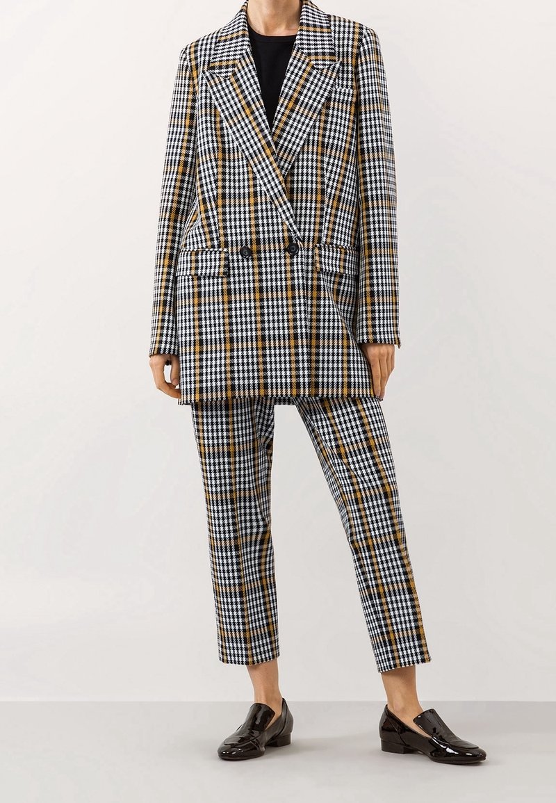 Personne portant un blazer croisé à carreaux noir, blanc et jaune moutarde avec un pantalon court assorti et des mocassins noirs brillants.