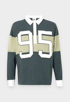 Μακρυμάνικο πόλο σε σκούρο πράσινο και μπεζ, με λευκό γιακά και μεγάλο λευκό αριθμό "95" στο μπροστινό μέρος. Υφή από μείγμα βαμβακιού.