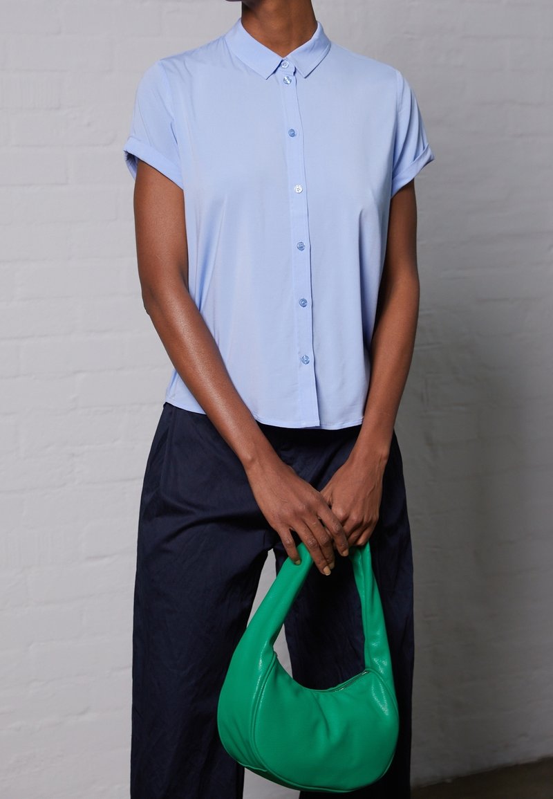Personne portant une chemise bleu clair à manches courtes boutonnée et un pantalon foncé tenant un sac à main vert vif devant un mur de briques gris.