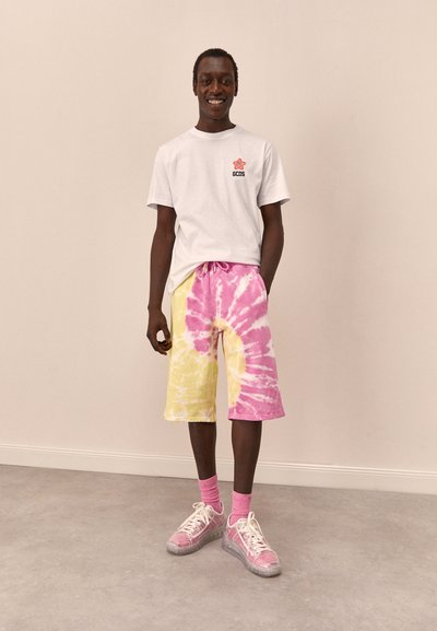 Fehér pamut póló piros logóval, rózsaszín és sárga batikolt rövidnadrággal párosítva. Az öltözéket rózsaszín zoknik és színes tornacipők egészítik ki.