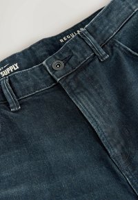 Jeans din denim albastru închis, cu o croială regulară, prezentând o închidere cu nasture, cusături proeminente și un finisaj texturat al țesăturii.