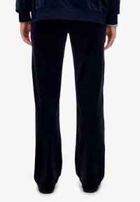 Persoon in donker marineblauwe, losse broek en een bijpassende marineblauwe sweatshirt, staande tegen een effen witte achtergrond.