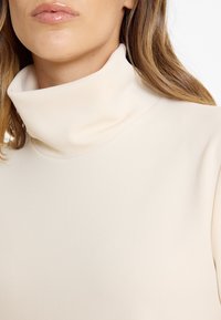 Frau trägt ein Creme-Oberteil mit hohem Kragen und langen Ärmeln sowie glänzendem, hellrosa Lippenbalsam, zeigt Untergesicht und Hals.