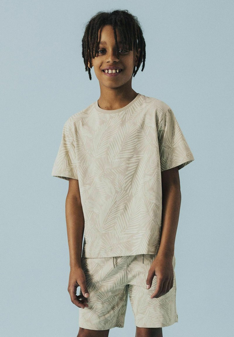 Short-sleeve beige shirt with a subtle leaf pattern, paired with matching shorts. Both items feature a relaxed fit and a soft texture.
