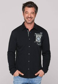 Camisa negra de manga larga con cuello, que presenta cierres de botón, estampados decorativos y una tela texturizada. Usada de manera casual con denim claro.