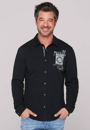 Camisa negra de manga larga con cuello, que presenta cierres de botón, estampados decorativos y una tela texturizada. Usada de manera casual con denim claro.