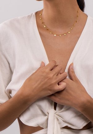 Femme portant un haut blanc à nouer sur le devant et un délicat collier en or avec de petits disques pendants, mains jointes près de sa poitrine.