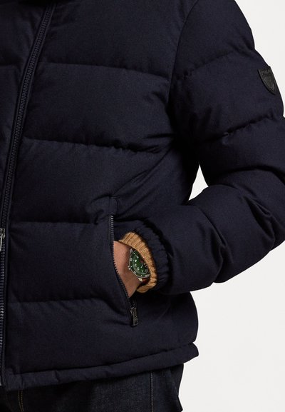 Navy puffer jacket with quilted pattern, zip closure, and ribbed cuffs. Features a logo patch on the shoulder and a pocket with a watch visible.