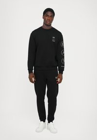 Black sweatshirt and sweatpants made of a soft fabric, featuring logos on the chest and sleeve, paired with white sneakers. Simple design.