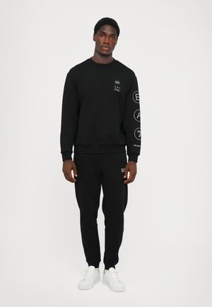 Black sweatshirt and sweatpants made of a soft fabric, featuring logos on the chest and sleeve, paired with white sneakers. Simple design.