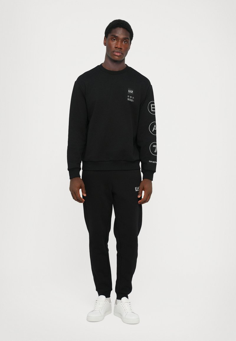 Black sweatshirt and sweatpants made of a soft fabric, featuring logos on the chest and sleeve, paired with white sneakers. Simple design.