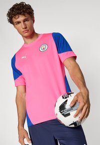 Ροζ ποδοσφαιρική φανέλα με μπλε μανίκια, με κυκλικό λογότυπο και άσπρη επωνυμία Puma. Ο μοντέλο κρατά μια ποδοσφαιρική μπάλα με ασπρόμαυρο σχέδιο.