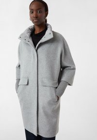 Grauer Wollmantel mit hohem Kragen, Druckknöpfen und zwei Fronttaschen. Mit gerippten Bündchen, die aus den Ärmeln hervorschauen, für zusätzliche Textur.