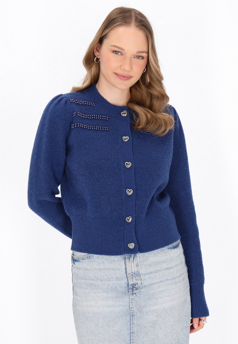 Cardigan lavorato a maglia blu con bottoni a forma di cuore, polsini a coste e decorazioni in perline sulle spalle. Texture morbida, design aderente.