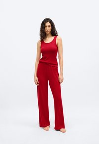 Γυναίκα με κυματιστά μαλλιά, ντυμένη με κόκκινο αμάνικο τοπ ριπ και ίσια παντελόνια με φαρδιά πόδια, όρθια ξυπόλητη σε απλό ανοιχτό φόντο.