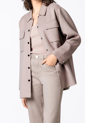 Frau trägt eine beige Wolljacke mit Knöpfen über einem gerippten Tanktop, die Hand in der Tasche beige passender, hochgeschnittener Hose.