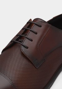 Κοντινή εικόνα καφέ δερμάτινου παπουτσιού για επίσημες εμφανίσεις με διάτρητη μύτη, λείο πέλμα και μαύρα κορδόνια, σε λευκό φόντο.