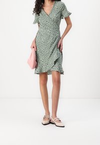 Grön wrapklänning med korta volangärmar, svarta bladmönster och utställd fåll. Matchad med en rosa väska och beige skor med accenter.