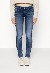 Jeans ajustados de mezclilla azul con una cintura de altura media, ligero desgaste y detalles de costura sutiles. Combinados con zapatos blancos con cordones coloridos.