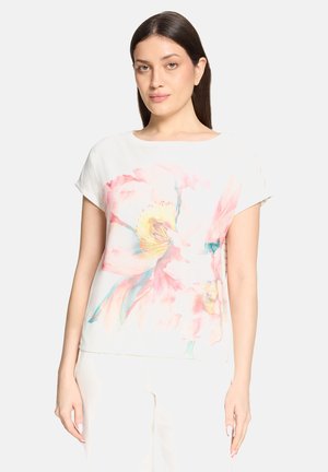 Kobieta o długich ciemnych włosach, ubrana w białą bluzkę z krótkim rękawem, zdobioną dużym pastelowym nadrukiem kwiatowym w odcieniach różu i żółci.