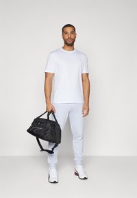 White short-sleeve t-shirt, light gray sweatpants, white sneakers with black accents, holding a black duffel bag with a white logo.