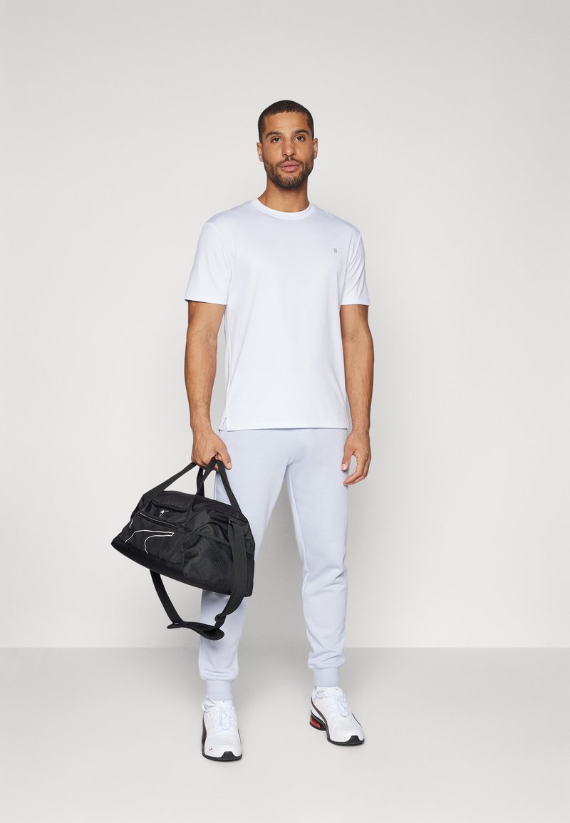 Camiseta blanca de manga corta, pantalones de chándal gris claro, zapatillas blancas con detalles negros, sosteniendo un bolso de deporte negro con un logo blanco.
