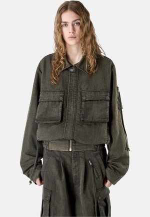 Jonge persoon met lang golvend haar, gekleed in een oversized donkerolijfgroene jas met grote voorzakken en bijpassende cargobroek.