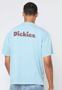 Camiseta de algodón azul claro con mangas cortas. Logotipo de "Dickies" en marrón situado de manera destacada en la parte trasera. Cuello redondo y corte holgado.