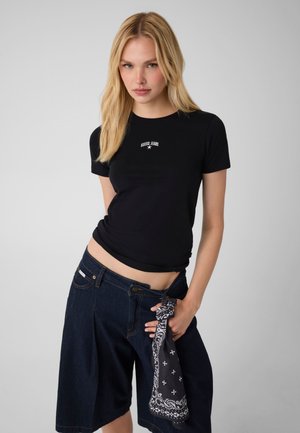 Giovane donna con capelli biondi che indossa una t-shirt nera Guess Jeans e pantaloncini di denim scuri, che tiene in mano una bandana nera con fantasia.