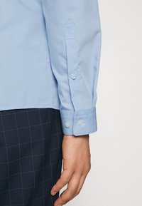 Lichtblauwe blouse met lange mouwen, met knoopmanchetten en een spitse kraag, gemaakt van gladde stof, gecombineerd met donkere geruite broek.