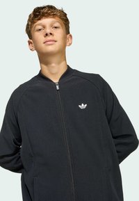 Black zip-up jacket made of ribbed fabric, featuring a stand-up collar and discreet white logo on the left chest.